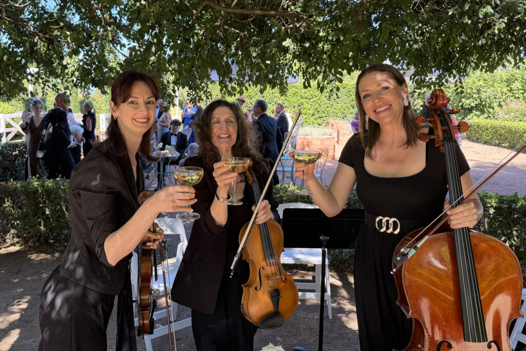 Professional string trio performing live at an outdoor garden wedding in Sydney. Three female musicians playing violin, viola, and cello during a wedding ceremony.