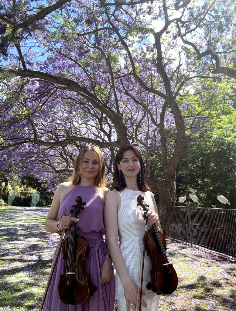 Two female violinists holding their instruments, posing beneath a vibrant, blooming purple jacaranda tree.