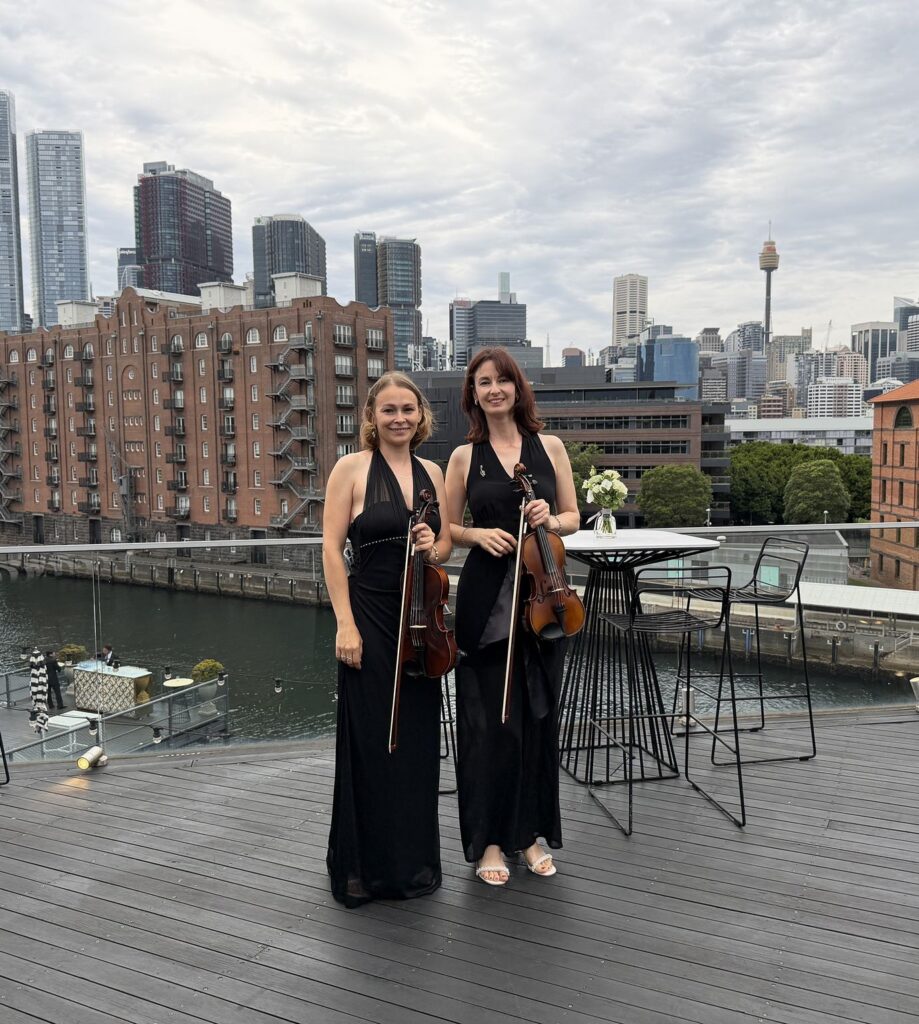 Two professional female violinists in black formal attire standing on a rooftop balcony overlooking the Sydney CBD skyline and Harbour.