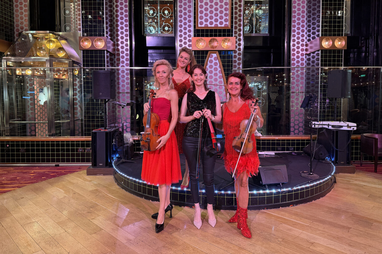 A professional quartet of female violinists performing on a stage with a mix of electric violins and classic violins. Three musicians in red dresses and one in black, perfect for high-energy corporate event entertainment in Sydney.
