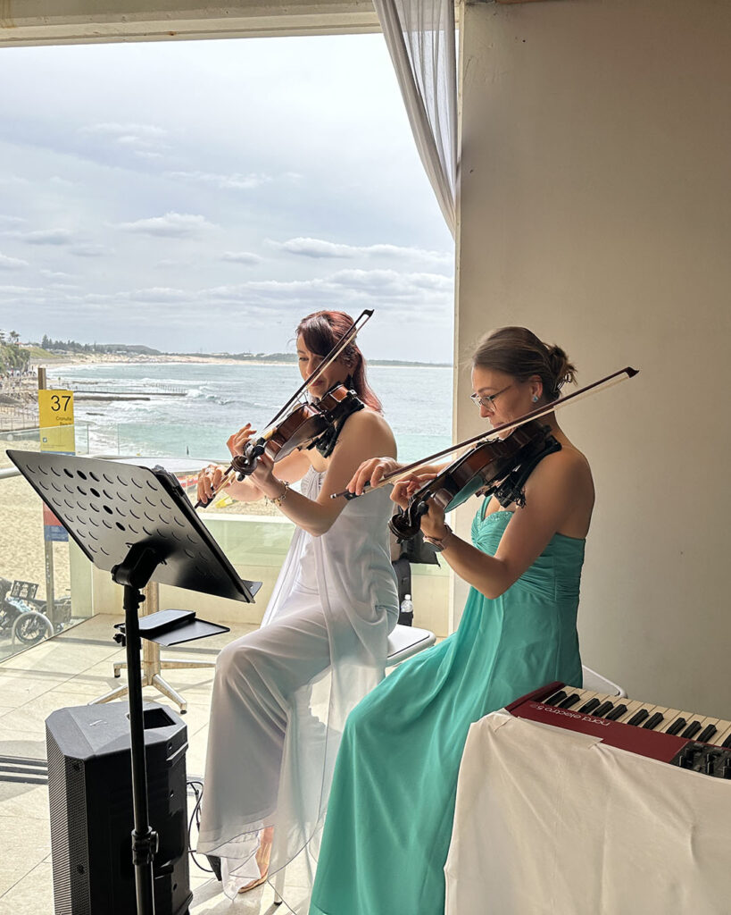 Two elegant female violinists of Luminous Strings performing live. One is in a silver dress with a music stand, and the second is in a teal dress, positioned next to a keyboard.