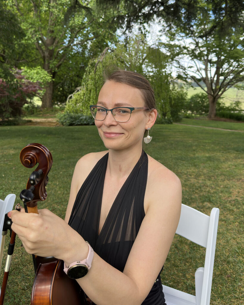 A professional female violinist in a black formal dress holding a violin at an outdoor garden wedding venue.
