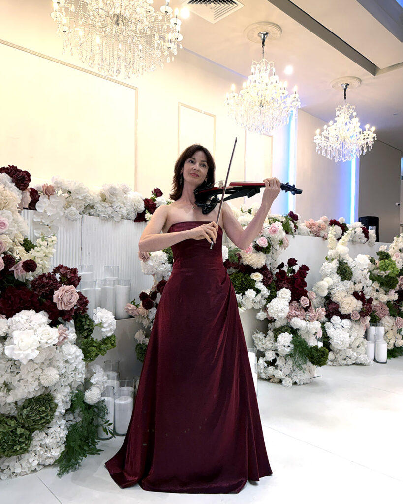 Professional female electric violinist in a burgundy dress performing solo live music on stage at a modern wedding ceremony with floral backdrop.