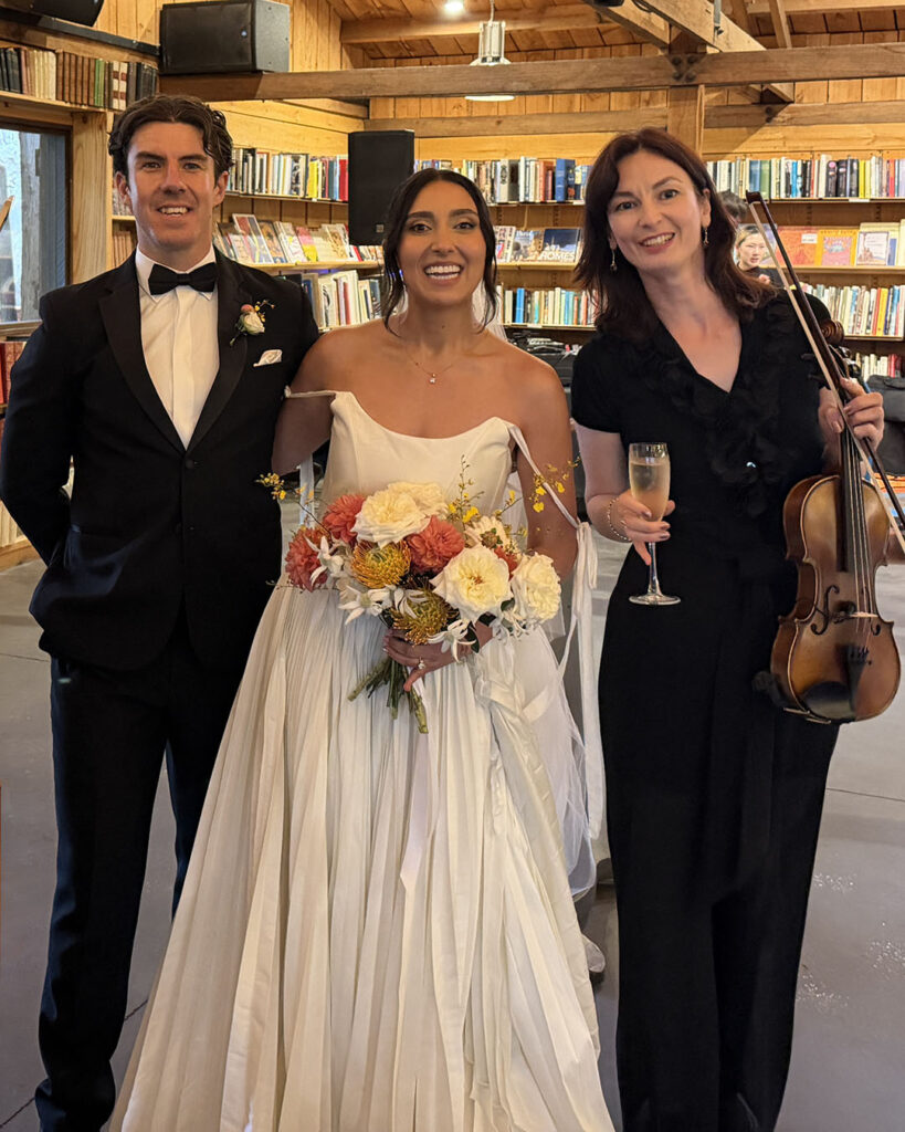 A happy bride and groom posing with a professional female violinist at their wedding reception venue.