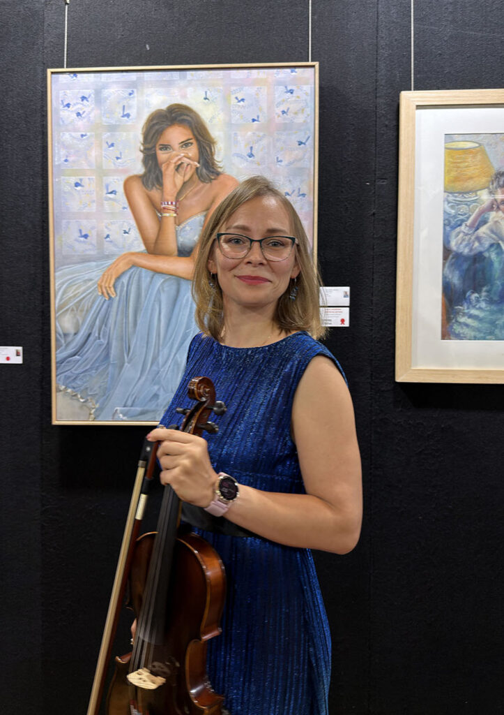 A blonde woman in a royal blue sleeveless dress holding a violin, standing in front of framed paintings at an art exhibition center.