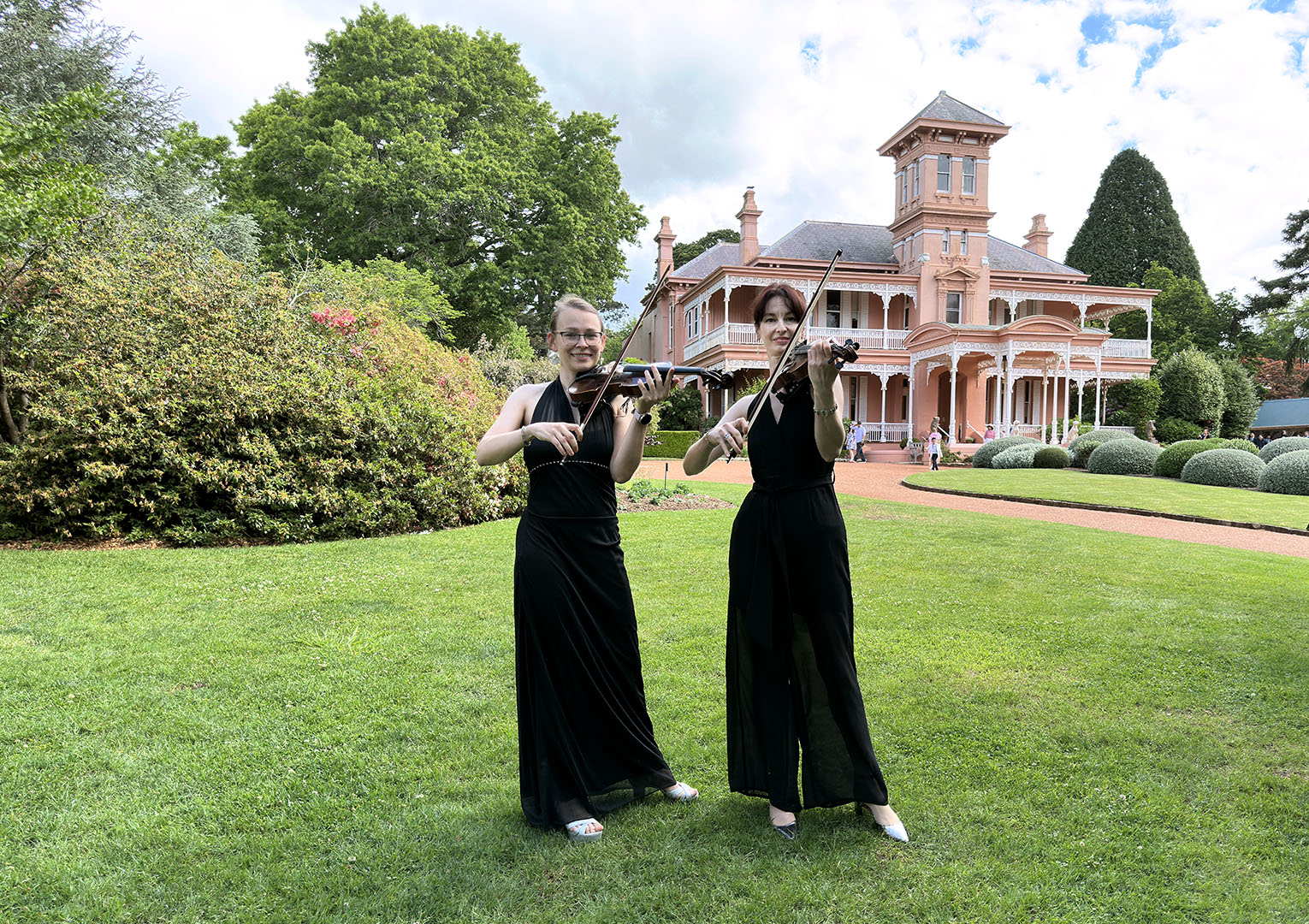 Two professional female violinists in black formal dresses performing as a string duo at a luxury estate garden wedding.