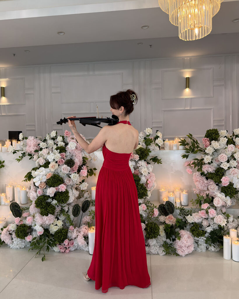 Female electric violinist in a red formal dress performing a solo at an event with floral decorations.