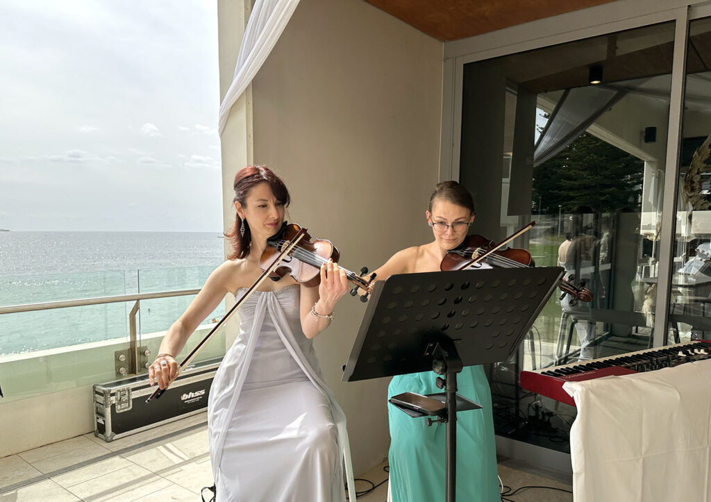 Two professional wedding violinists from Luminous Strings performing live as a duo during an outdoor oceanfront wedding ceremony.