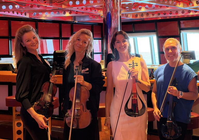 Four professional wedding violinists holding their instruments, smiling together at an event venue.
