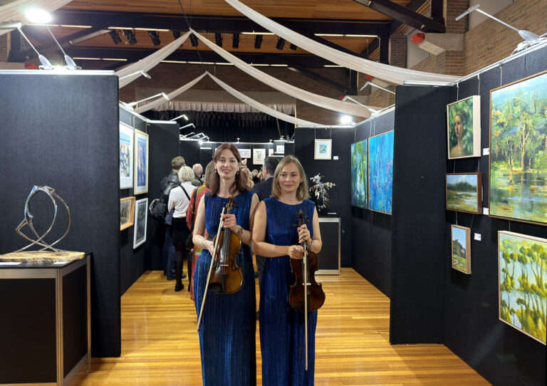 Two professional female violinists from Luminous Strings in elegant blue dresses, holding their instruments at a Sydney corporate event and art exhibition.
