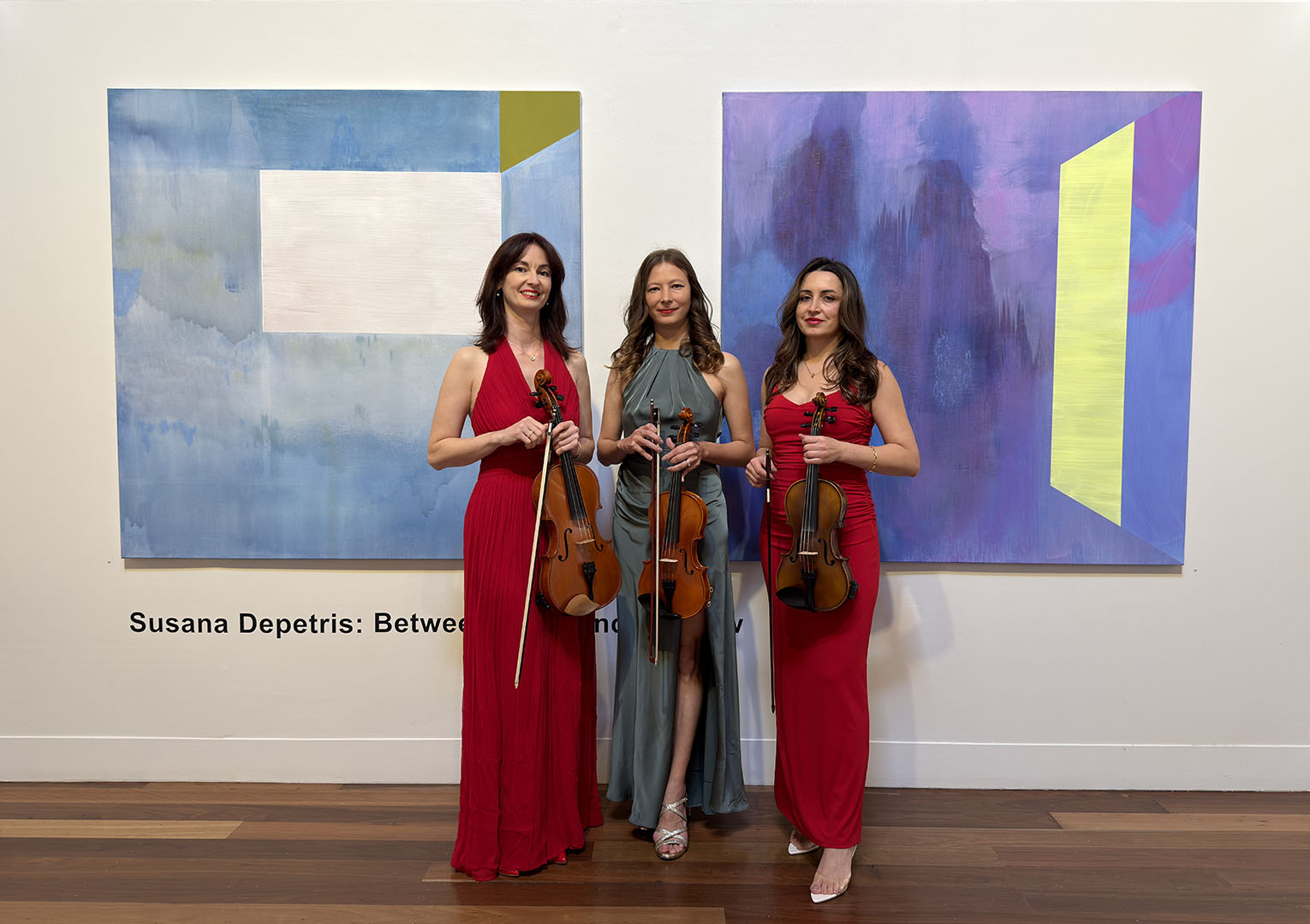 Three elegant female violinists in formal red and grey evening gowns holding their violins, standing in an art gallery for a live performance event.