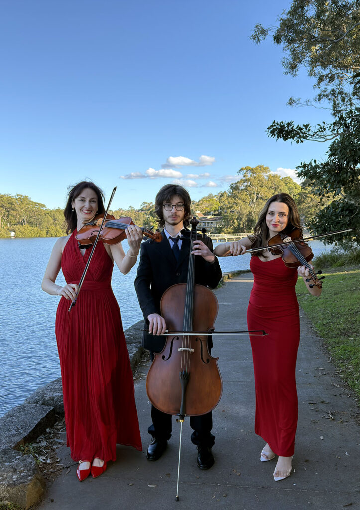 A classical string trio featuring a cellist seated in a suit and two female violinists/violists standing in matching red dresses, performing outdoors near a lake.