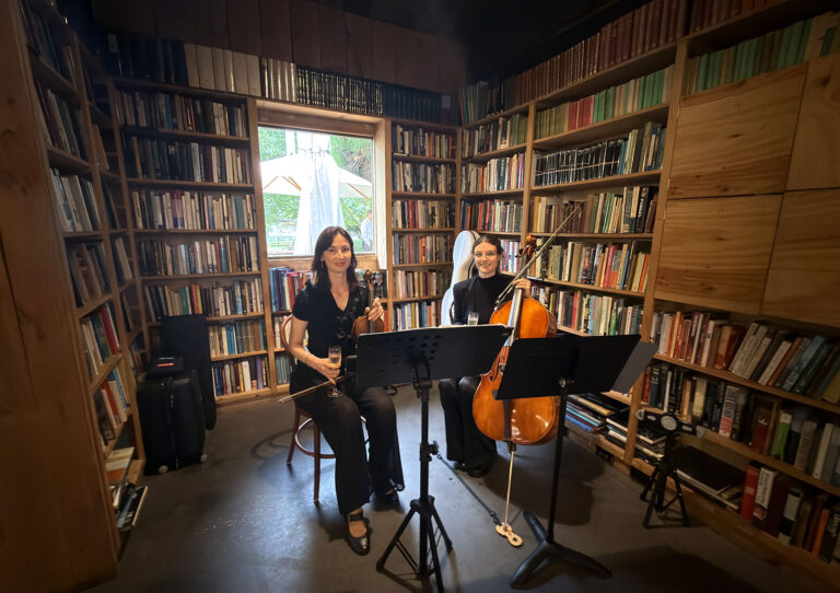 A classical string duo featuring a female violinist and a cellist performing with music stands in a cozy, library-style room for a birthday party.