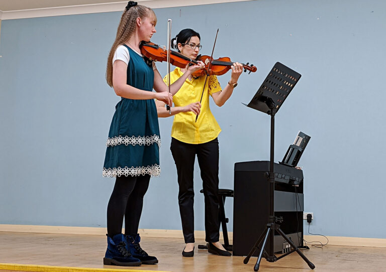 Two female classic string duo musicians playing violins next to a digital piano on a stage during a birthday party event performance.
