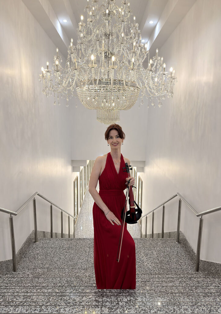 A female solo violinist in a formal red evening gown holding a violin under a large crystal chandelier at a birthday party function hall.