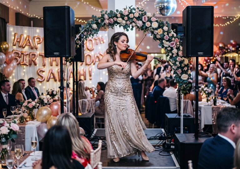 A female solo violinist in a formal evening gown performing live in front of a floral arch and audio speakers at a birthday party reception.