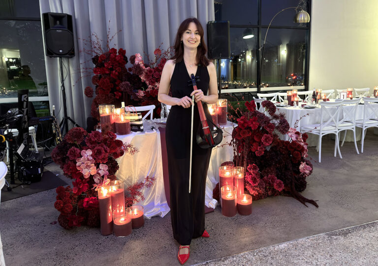 A female solo violinist standing with a wireless electric violin in front of elegant red floral decor at an evening birthday party event.