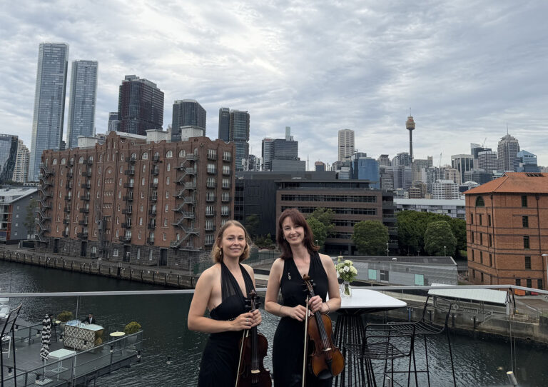 Two female string duo musicians holding violins while posing outdoors for a birthday party event with a city skyline in the background.