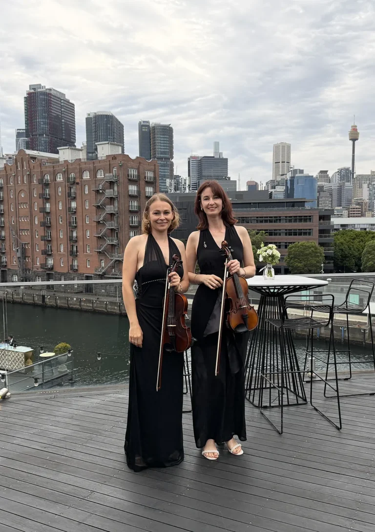 Two professional female violinists in black formal attire standing on a rooftop balcony overlooking the Sydney CBD skyline and Harbour.