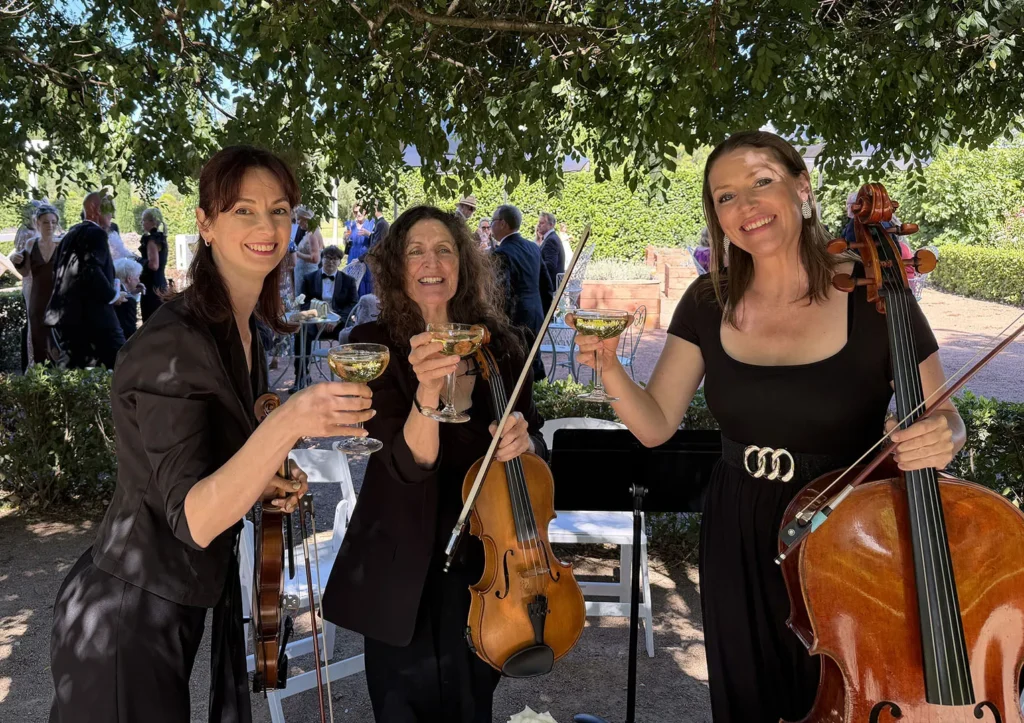 Professional string trio performing live at an outdoor garden wedding in Sydney. Three female musicians playing violin, viola, and cello during a wedding ceremony.
