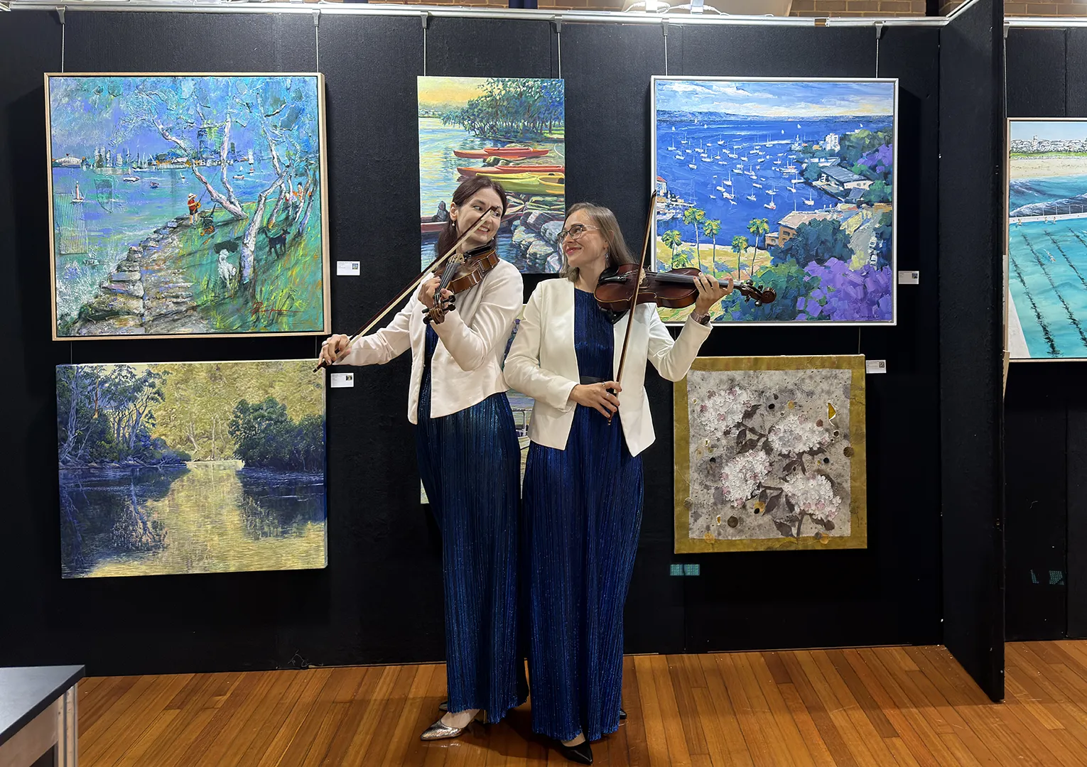 Two female violinists in white jackets playing violins together inside an art gallery for a PlanEvent performance.