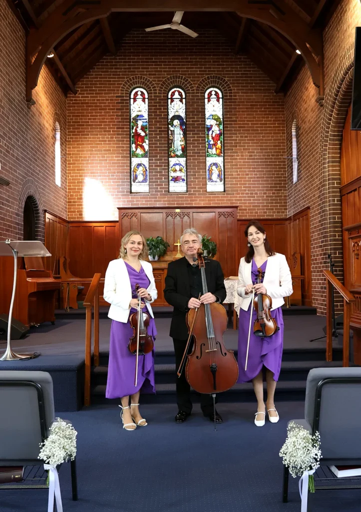String trio performing at a church ceremony, featuring two female violinists and a male cellist.