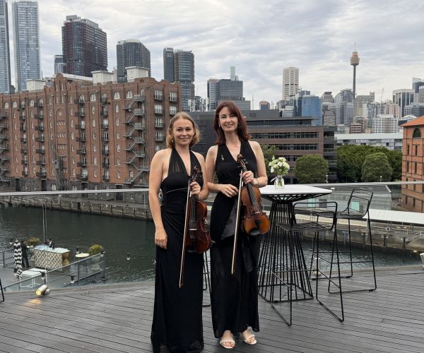 Two professional female violinists in black formal attire standing on a rooftop balcony overlooking the Sydney CBD skyline and Harbour.